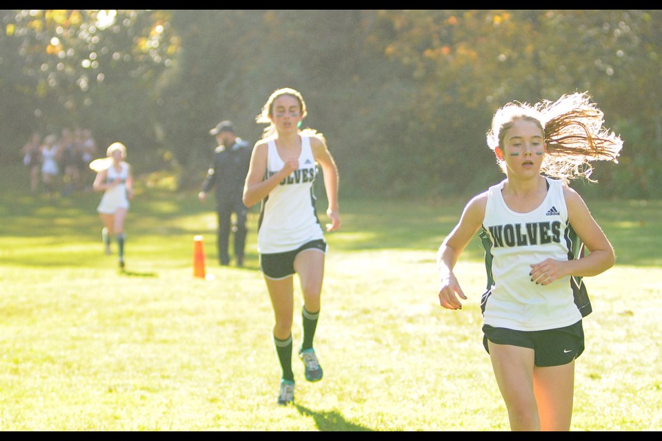 Audrey Warner streaks across a meadow at Jericho Park with Sophie Dodd and Leiah Kirsh behind her. West Point Grey Academy coach Sean Dawson stands beside the course in the distance.