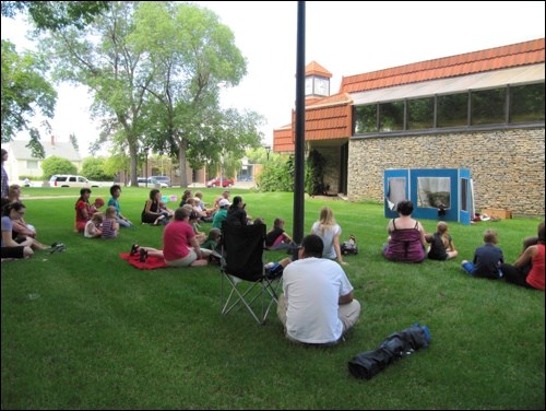 In the park behind the Library the kids and parents are sitting and enjoying the puppet show by the Fort Battleford junior volunteers.