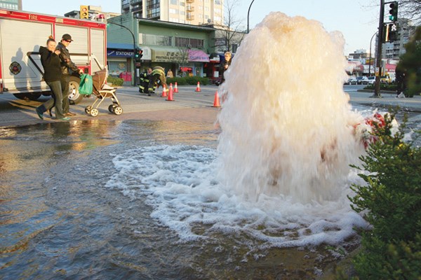 As firefighters work to turn off a water valve, passing pedestrians take photos and watch as an overturned fire hydrant spews water into the intersection of 16th St W and Lonsdale Ave.