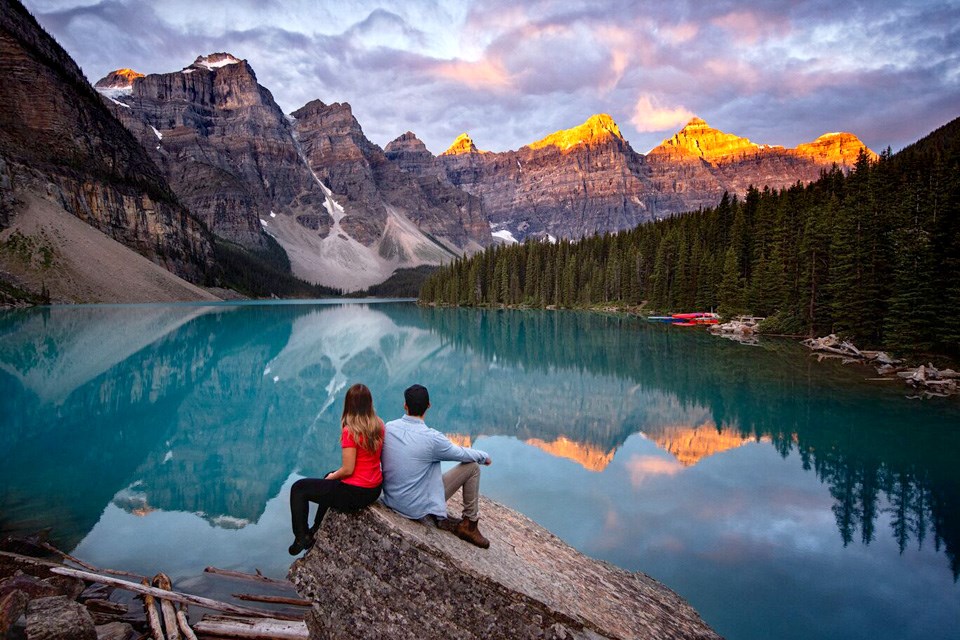 Moraine Lake: A woman and a man sit on a rock and look out over Moarine Lake as the sun hits the mountain peaks / Banff & Lake Louise Tourism