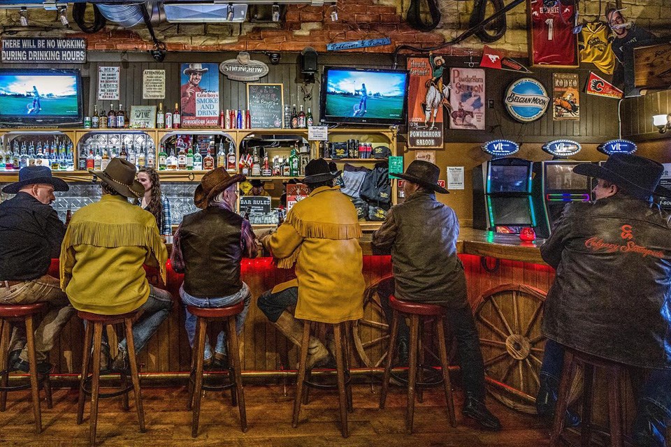 Six men in cowboy hats and jackets from behind as they sit at the bar of The Powderhorn Saloon in Alberta / The Powderhorn Saloon