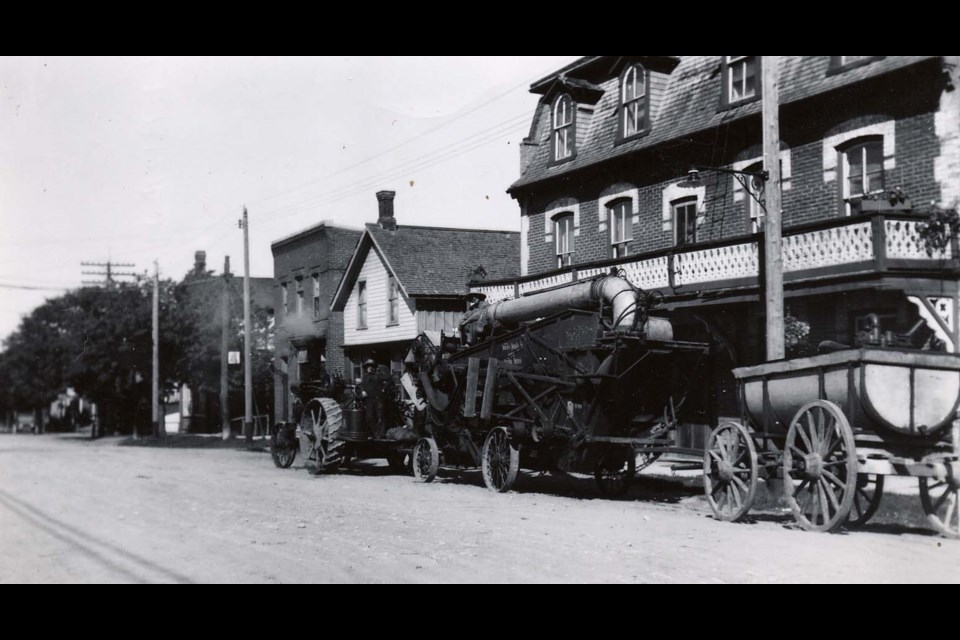 Photo of downtown Cookstown circa 1900 taken of the north side of Queen Street. The buildings visible in the image from left to right are the butcher shop, telephone office, Eby's Store (also known as Bob Robinson's), and the Mansion House Hotel. The image also features some large machinery, a threshing machine and water tank owned by the Water Ferrier of Innisfil, parked in front of the Mansion House Hotel being towed by a tractor and operated by two men. The telephone lines are also visible, but the image predates the installation of hydro lines.