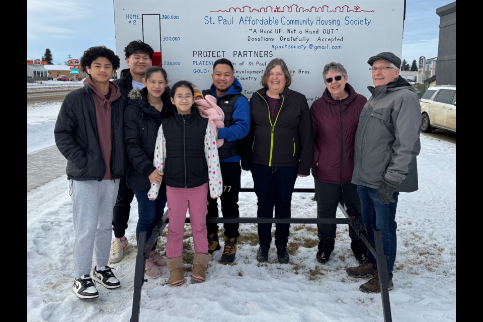 (Left to right) Kirt, Kyrone, Menchu, Cassandra, Jay-ar, and Queen Levin Guardiana, Louise Severin, Alice Bourget, and Guy Laberge in front of the St. Paul Affordable Community Housing Society fundraising sign on Main Street St. Paul. The Guardiana family was selected for an affordable home, which the society expects to break ground on in spring 2025. 