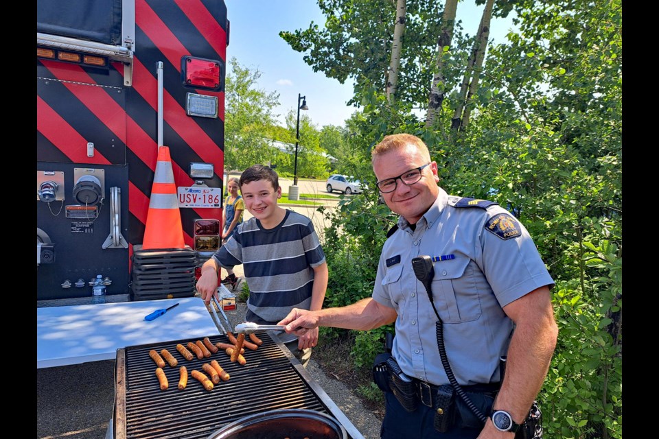 Innisfail Staff Sgt. Ian Ihme, the detachment commander, gets assistance at cooking hotdogs at the annual Bike Rodeo at the Innisfail Library/Learning Centre on June 28. Submitted photo