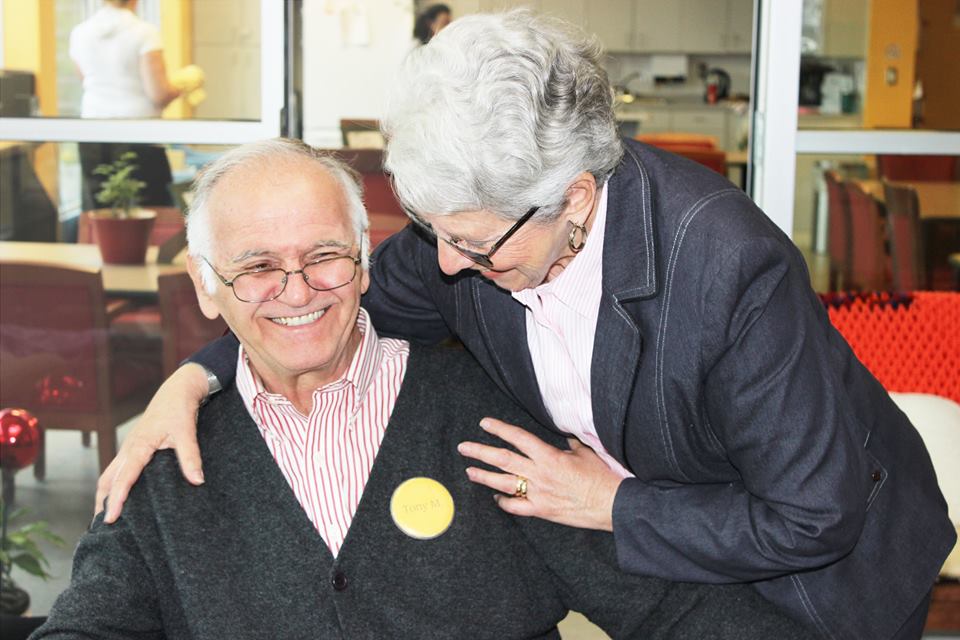 Elderly couple indoors with wife hugging husband who is sitting down | United Way Oakville