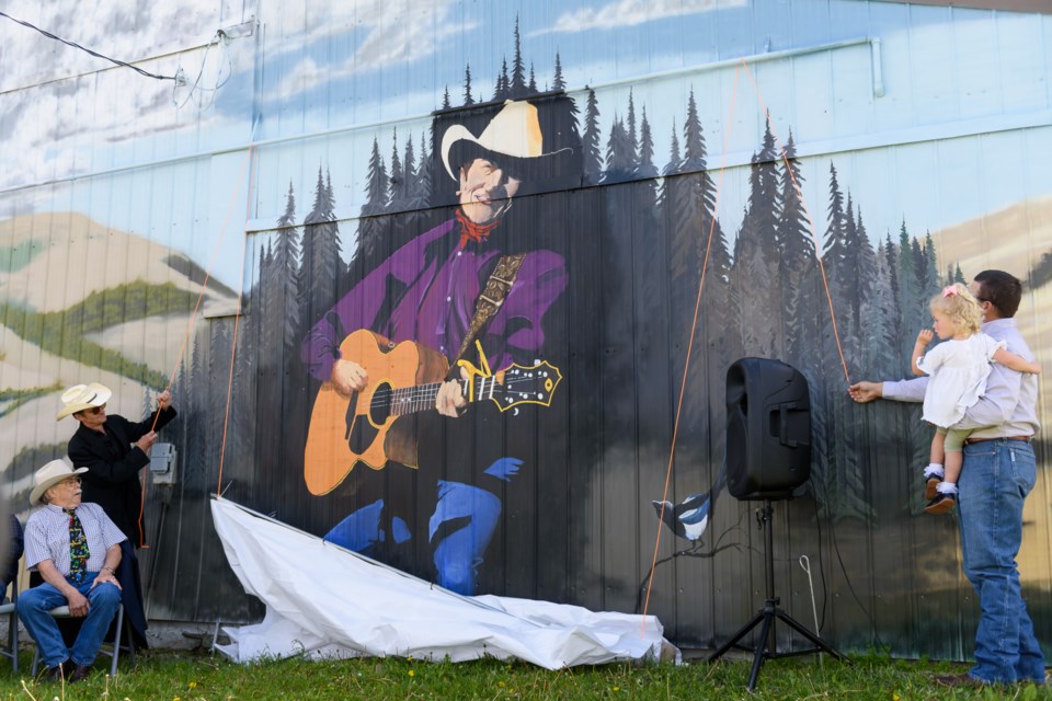 Ian Tyson’s son Clay Tyson, left, wearing black,  lowers the curtain to unveil a mural of Ian Tyson with Ian's son-in-law, Hunter Bell, and Bell's daughter Mesa, right, at  East Longview Hall in Foothills County on July 4. 