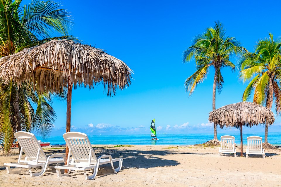 Sun loungers under straw umbrellas on the sandy beach with palms near ocean with sailboat. Vacation background. Idyllic beach landscape. Varadero, Cuba.